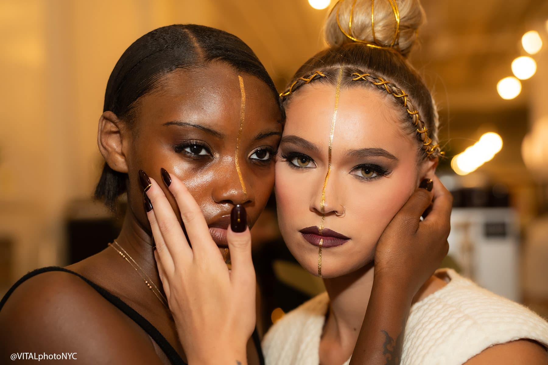 Two models cheek to cheek backstage, each with a painted line down the face