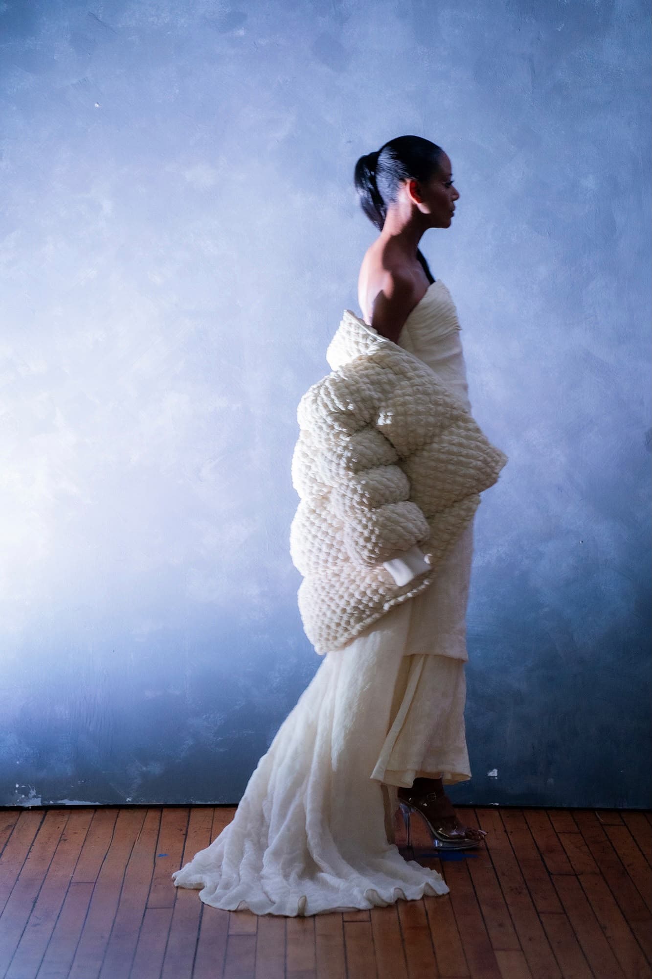 A cream quilted jacket worn off the shoulders over a long white dress, photographed in profile