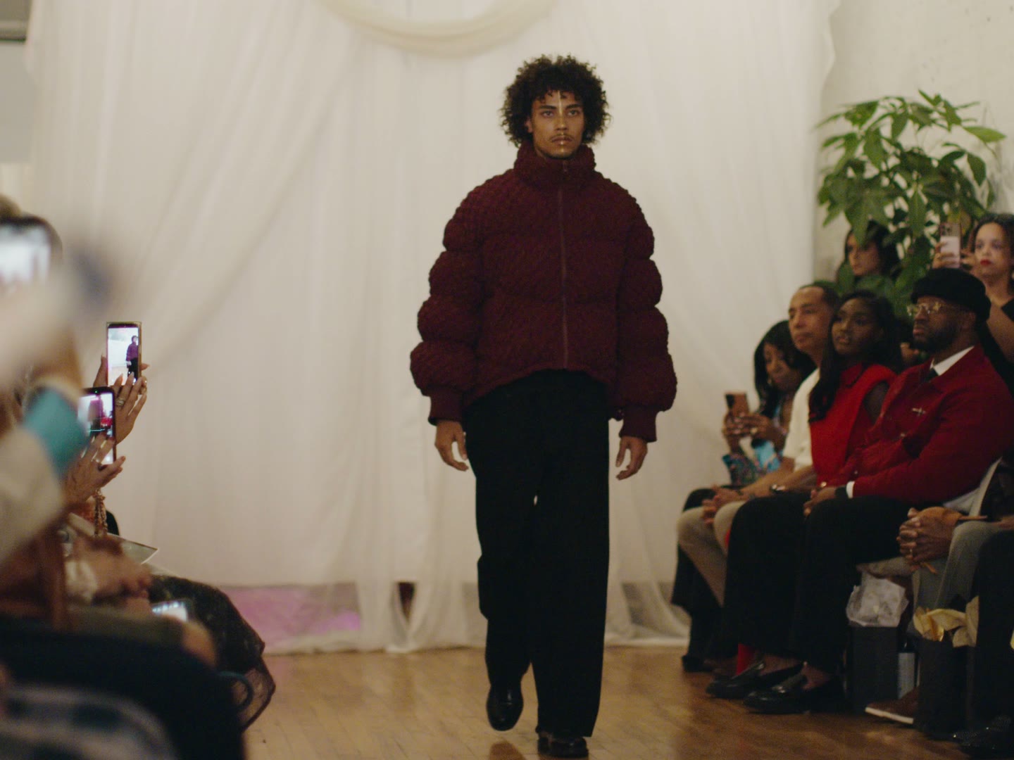 A model in a burgundy quilted puffer and black trousers walks the Between Worlds runway between seated guests